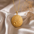 Gold Plated Silver Lunar Impact Pendant with a crater-textured round disc and radiating lines on champagne satin beside dried flowers