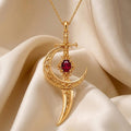 Gold Plated Silver Moonblade pendant with an openwork crescent, cross-shaped upper detail, red marquise-shaped center stone, and tapered lower drop on champagne satin